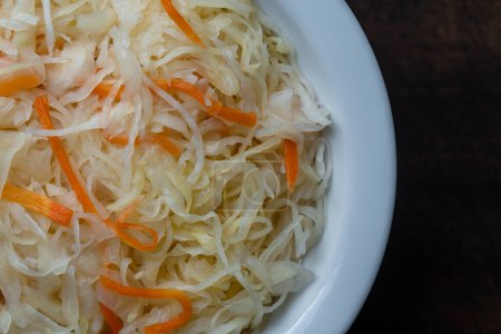 Photo for Homemade sauerkraut in ceramic bowl on a wooden background, close up, top view. Finely cut cabbage with carrots, fermented by lactic acid bacteria with long shelf life and distinctive sour flavor - Royalty Free Image