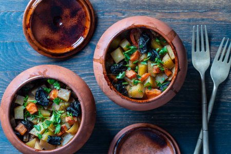 Photo for Stewed potato, carrot, onion, tomato and prunes in a clay pots with lid on wooden background, close up, top view - Royalty Free Image