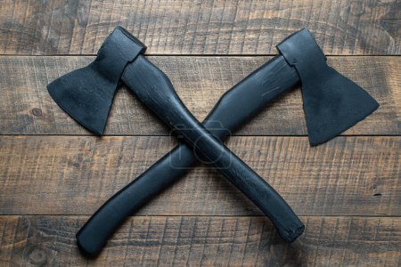 Two crossed black axes on wooden background, top view, close up