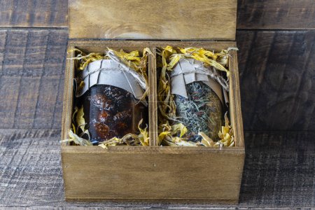 Photo for Glass jar with of flower tea and a glass jar with of pine cone jam in a wooden gift box on a table, close up. Beautiful present for family and friends - Royalty Free Image