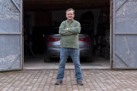 Foto de Sonriente hombre adulto mecánico se para con los brazos cruzados delante de un garaje de coches, de cerca - Imagen libre de derechos