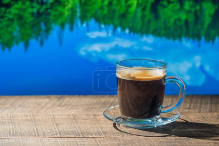 Foto de Taza de vidrio de café negro caliente en la mesa de madera con el lago y el fondo del bosque en un día soleado de verano, de cerca - Imagen libre de derechos