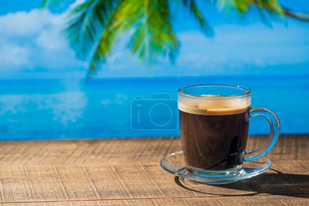 Foto de Taza de vidrio de café negro caliente en mesa de madera con agua de mar, palmera de coco y fondo de cielo azul en el soleado día de verano en el café de playa tropical, de cerca, espacio para copiar - Imagen libre de derechos