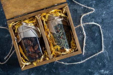 Foto de Frasco de vidrio con de té de flores y un frasco de vidrio con de mermelada de cono de pino en una caja de regalo de madera en una mesa, de cerca. Hermoso regalo para la familia y amigos - Imagen libre de derechos
