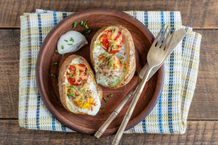 Foto de Patatas al horno rellenas de queso, tomate, cebolla verde y huevos en plato de cerámica sobre fondo de madera, vista superior, primer plano - Imagen libre de derechos