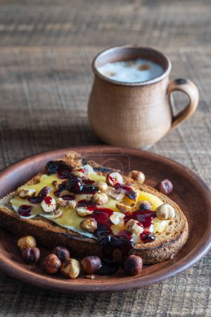 Foto de Pan tostado con mantequilla, avellanas asadas, miel, mermelada de bayas en el plato y café capuchino en una taza de arcilla en la mesa de madera, de cerca - Imagen libre de derechos