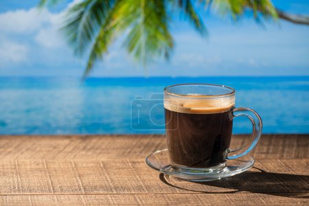 Foto de Taza de vidrio de café negro caliente en mesa de madera con agua de mar, palmera de coco y fondo de cielo azul en el soleado día de verano en el café de playa tropical, de cerca, espacio para copiar - Imagen libre de derechos