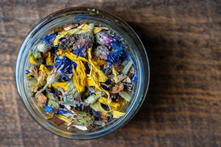 Foto de Dry flower and herbal tea leaves in a glass jar on wooden background, copy space. Herbal collection of chamomile, cornflower, mint, sea buckthorn, lemongrass, wild rose, dried citrus fruits and apple - Imagen libre de derechos