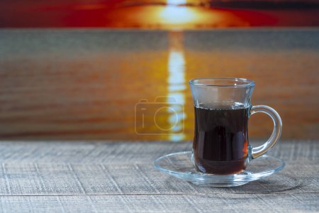 Foto de Taza de vidrio de té negro caliente en la mesa de madera con agua de mar durante el atardecer en el día de verano en el café tropical de la playa, de cerca, espacio de copia - Imagen libre de derechos