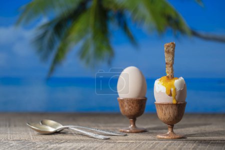 Photo for Soft boiled egg in eggcup with slice of toasted toast on wooden table with sea water, coconut palm tree and blue sky background on sunny summer day in tropical beach cafe, close up - Royalty Free Image