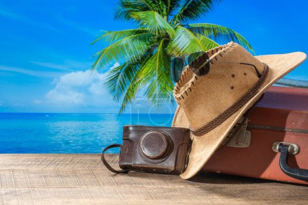 Téléchargez les photos : Valise, chapeau de soleil, appareil photo et lunettes de soleil sur le pont en bois avec de l'eau de mer, cocotier et fond bleu ciel sur une journée ensoleillée d'été à la plage tropicale, espace de copie. Concept vacances et voyages - en image libre de droit