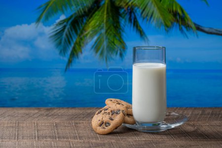 Foto de Leche blanca fresca en un vaso con galletas en una mesa de madera con agua de mar, palmera de coco y fondo azul en el soleado día de verano en la cafetería tropical de playa, de cerca - Imagen libre de derechos