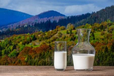 Frische weiße Milch in einem Krug und Glas auf einem Holztisch vor dem Hintergrund der herbstlichen Karpaten, aus nächster Nähe. Milchproduktekonzept