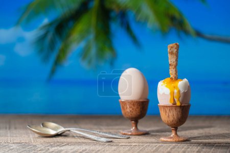 Photo for Soft boiled egg in eggcup with slice of toasted toast on wooden table with sea water, coconut palm tree and blue sky background on sunny summer day in tropical beach cafe, close up - Royalty Free Image