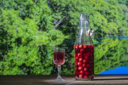 Foto de Brandy de cereza hecho en casa en una copa de cristal y una botella de vidrio en una mesa de madera sobre fondo natural en un día soleado de verano, de cerca - Imagen libre de derechos
