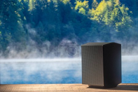 Photo for Black acoustic sound speaker on a wooden table with the lake water and forest background on a sunny summer day. The musical equipment, close up - Royalty Free Image