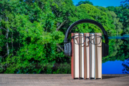 Photo for Set of books, headphones and eyeglasses on wooden table with the lake water and forest background on a sunny summer day, close up. Audiobooks and education concept - Royalty Free Image