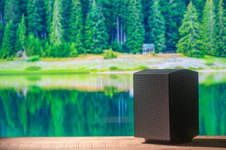 Téléchargez les photos : Haut-parleur acoustique noir sur une table en bois avec l'eau du lac et le fond de la forêt par une journée d'été ensoleillée. L'équipement musical, gros plan - en image libre de droit