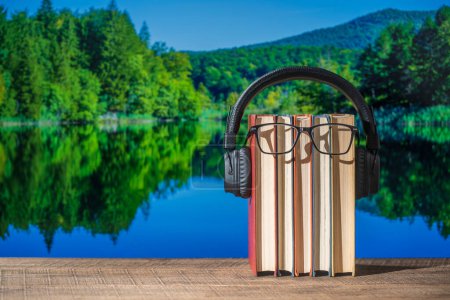Téléchargez les photos : Ensemble de livres, écouteurs et lunettes sur une table en bois avec l'eau du lac et le fond de la forêt par une journée d'été ensoleillée, fermer. Livres audio et concept d'éducation - en image libre de droit