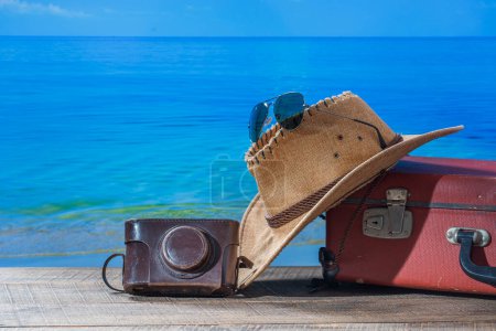 Téléchargez les photos : Valise, chapeau de soleil, appareil photo et lunettes de soleil sur le pont en bois avec fond d'eau de mer par une journée ensoleillée d'été à la plage tropicale, espace de copie. Concept vacances et voyages - en image libre de droit