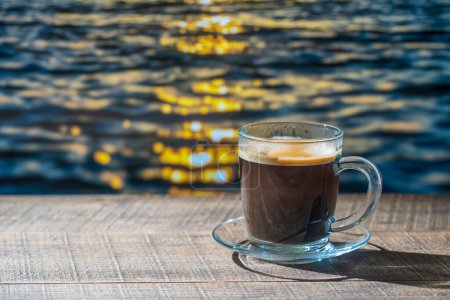 Foto de Taza de vidrio de café negro caliente en mesa de madera con fondo de agua de mar en el día de verano en la cafetería tropical de la playa durante la puesta del sol, primer plano, espacio de copia - Imagen libre de derechos
