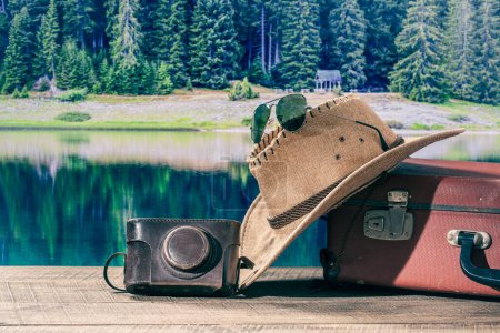 Téléchargez les photos : Valise, chapeau de soleil, appareil photo et lunettes de soleil avec l'eau du lac et le fond de la forêt par une journée d'été ensoleillée, fermer. Concept vacances et voyages - en image libre de droit