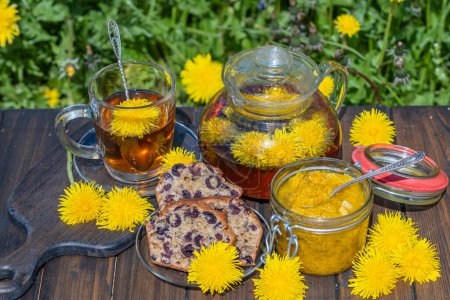 Foto de Té de flores de diente de león saludable en una tetera de vidrio en la mesa de madera junto con mermelada dulce y magdalena de cereza en el jardín de primavera, de cerca - Imagen libre de derechos