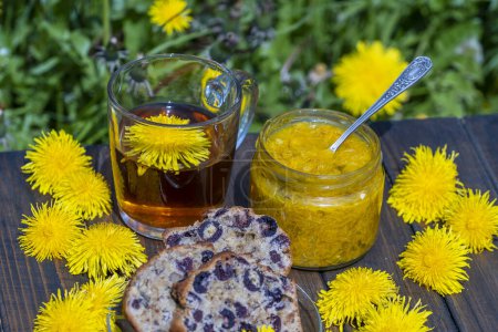Téléchargez les photos : Thé de fleur de pissenlit sain dans une tasse en verre sur la table en bois avec confiture douce et muffin de cerise dans le jardin de printemps, gros plan - en image libre de droit