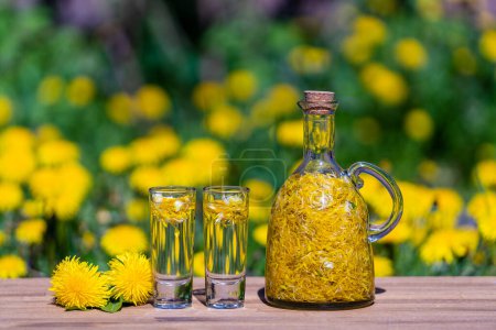 Foto de Diente de león hecho en casa flores tintura en dos vasos y en una botella de vidrio en una mesa de madera en un jardín de verano, de cerca - Imagen libre de derechos