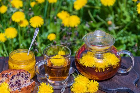 Téléchargez les photos : Thé de fleur de pissenlit sain dans une théière en verre sur la table en bois avec confiture douce et muffin de cerise dans le jardin de printemps, gros plan - en image libre de droit