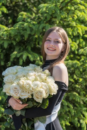 Téléchargez les photos : Portrait d'une belle fille heureuse avec un beau bouquet de roses blanches regardant la caméra et souriant avec charme, gros plan, à l'extérieur. Diplômé de l'école lors d'une cérémonie - en image libre de droit