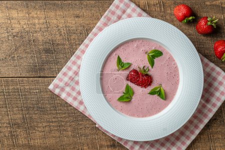 Sweet cold strawberry soup with green mint leaf in a white bowl on wooden table, close up, top view