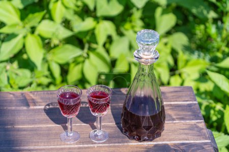 Photo for Homemade cherry brandy in glasses and in a glass bottle on a wooden table in a summer garden, close up - Royalty Free Image