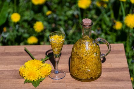 Foto de Tintura casera de flores de diente de león en un vaso de chupito y en una botella de vidrio sobre una mesa de madera en un jardín de verano, de cerca - Imagen libre de derechos