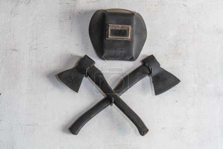 Foto de Dos hachas negras cruzadas y una máscara protectora para soldar en una pared de estuco blanco, de cerca - Imagen libre de derechos
