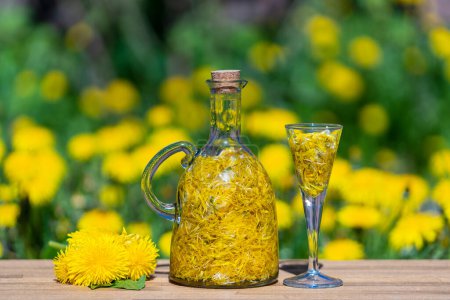 Photo for Homemade tincture of dandelion flowers in a shot glass and in a glass bottle on a wooden table in a summer garden, close up - Royalty Free Image