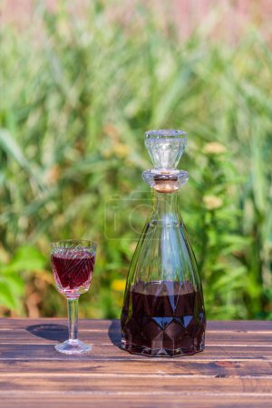 Téléchargez les photos : Homemade cherry brandy in glasses and in a glass bottle on a wooden table in a summer garden, close up - en image libre de droit