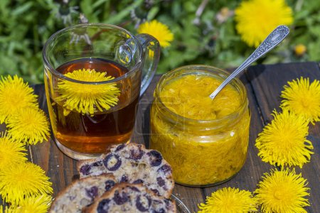 Photo for Healthy dandelion flower tea in a glass cup on the wooden table with sweet jam and cherry muffin in the spring garden, close up - Royalty Free Image