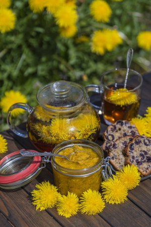 Téléchargez les photos : Thé de fleur de pissenlit sain dans une tasse en verre sur la table en bois avec confiture douce et muffin de cerise dans le jardin de printemps, gros plan - en image libre de droit