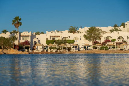 Téléchargez les photos : Bâtiments, chaises longues et parasols se reflètent dans l'eau de mer calme sur la plage dans la station balnéaire de Sharm El Sheikh pendant le lever du soleil, Egypte - en image libre de droit