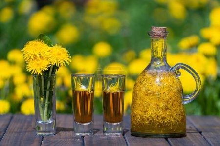 Foto de Diente de león hecho en casa flores tintura en dos vasos y en una botella de vidrio en una mesa de madera en un jardín de verano, de cerca - Imagen libre de derechos