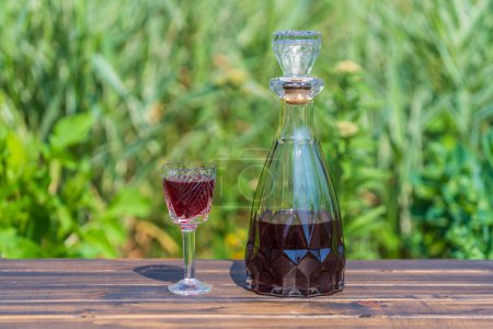 Téléchargez les photos : Homemade cherry brandy in glasses and in a glass bottle on a wooden table in a summer garden, close up - en image libre de droit