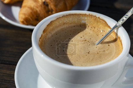 Foto de Espuma gruesa de capuchino café caliente con cuchara en taza blanca y croissant en mesa de madera, de cerca. Capuchino de espuma de leche, latte - Imagen libre de derechos