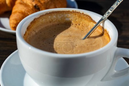 Téléchargez les photos : Mousse épaisse de cappuccino de café chaud avec cuillère en tasse blanche et croissant sur table en bois, fermer. Cappuccino en mousse de lait, latte - en image libre de droit