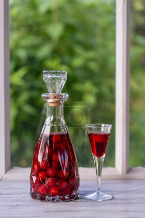 Foto de Brandy de cereza casero en copa de vino y en una botella de vidrio en un alféizar de ventana de madera cerca del jardín de verano, de cerca - Imagen libre de derechos