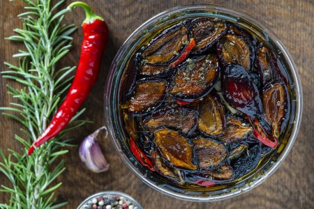 Sun-dried plums with garlic, green rosemary, red chili pepper, olive oil and spices in a glass jar on a wooden table. Rustic style, top view, close up