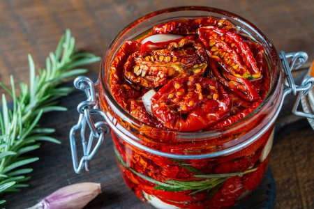 Photo for Sun-dried red tomatoes with garlic, green rosemary, olive oil and spices in a glass jar on a wooden table. Rustic style, close up - Royalty Free Image