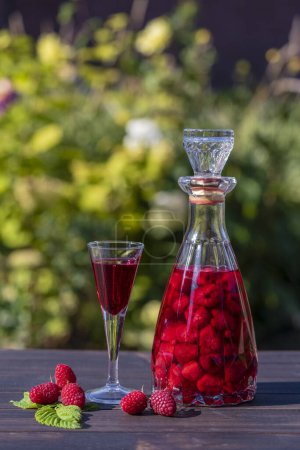 Photo for Homemade red raspberry brandy in glasses and in a glass bottle on a wooden table in a summer garden, close up - Royalty Free Image