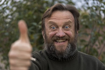 Photo for Portrait of cheerful optimist senior man looking at camera with thum up. Elderly bearded caucasian male in outdoor in urban park enjoying retirement, close up - Royalty Free Image