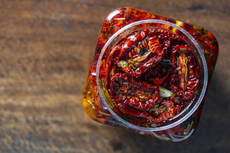 Photo for Sun-dried red tomatoes with garlic, green rosemary, olive oil and spices in a glass jar on a wooden table. Rustic style, top view, close up, copy space - Royalty Free Image